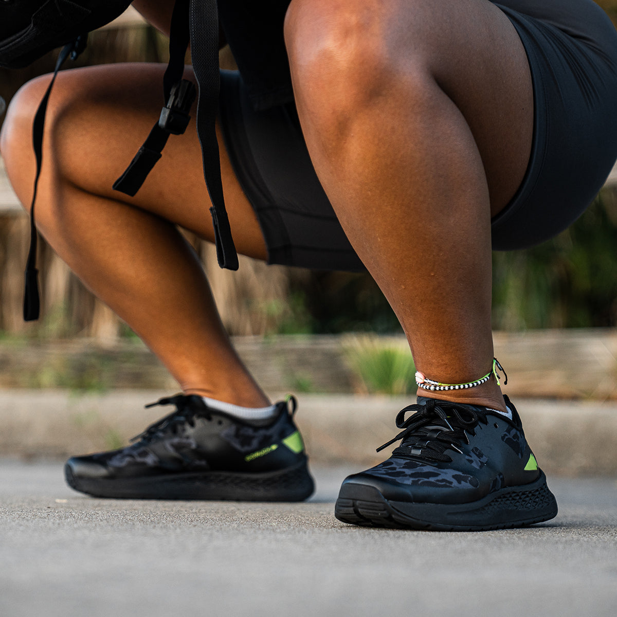 Wearing GORUCK's Women's Rough Runner - Midnight Frogskin + Acid Lime running shoes, a person squats on the pavement. They sport a silver ankle bracelet and black shorts, holding a black strap. The blurred wooden and grassy backdrop suggests they're gearing up for the Rough Runner challenge.