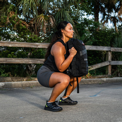 A person crouches in an outdoor setting, holding a black GORUCK backpack. Clad in Women's Rough Runner - Midnight Frogskin + Acid Lime athletic wear and lightweight running shoes, they appear engaged in exercise. Tall palm trees and a wooden fence form the backdrop, reminiscent of a Rough Runner adventure.