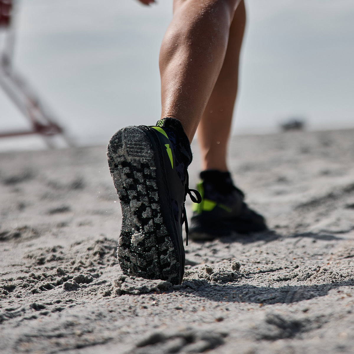 A person walks on a sandy beach, their legs and the sole of their Midnight Frogskin + Acid Lime Women's Rough Runner lightweight running shoes by GORUCK captured in focus. The sky is cloudy, and the sand appears coarse beneath the GRADIENT DENSITY EVA MIDSOLE.