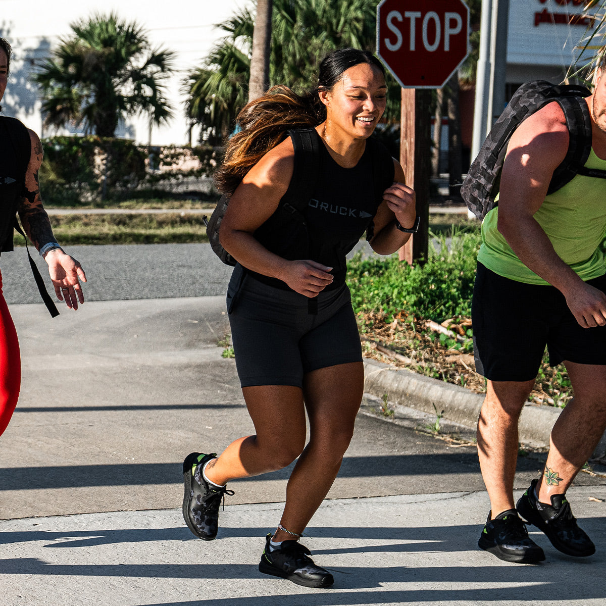 Three participants in Rough Runner are enjoying a sunny day, each donning athletic gear and backpacks. Their every stride is cushioned by the GRADIENT DENSITY EVA MIDSOLE in the Women's Rough Runner - Midnight Frogskin + Acid Lime by GORUCK. They move through a picturesque backdrop featuring a stop sign and palm trees.