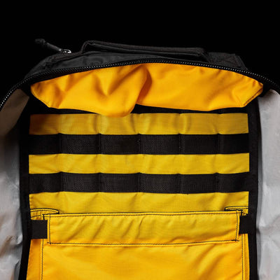 The GR1 USA - X-PAC by GORUCK showcases a partially open view, revealing its vibrant yellow interior with multiple horizontal black straps integrated into the lining. The exterior is crafted from durable black X-PAC fabric, with the zipper slightly visible at the top.