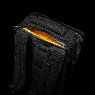 The GR1 USA - X-PAC by GORUCK is a black backpack featuring a quilted back panel and adjustable straps, partially open to reveal its bright orange interior against a dark background. Crafted from durable and waterproof X-PAC material, this backpack ensures both style and functionality.