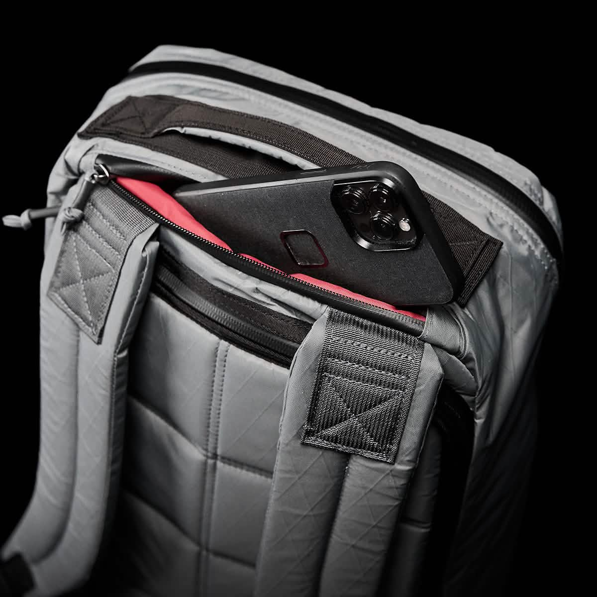 A sleek gray GORUCK GR1 USA - X-PAC rucksack featuring quilted straps and a textured design is partly unzipped, showcasing a smartphone with a black and red case nestled inside the pocket, all styled against an elegant black background.