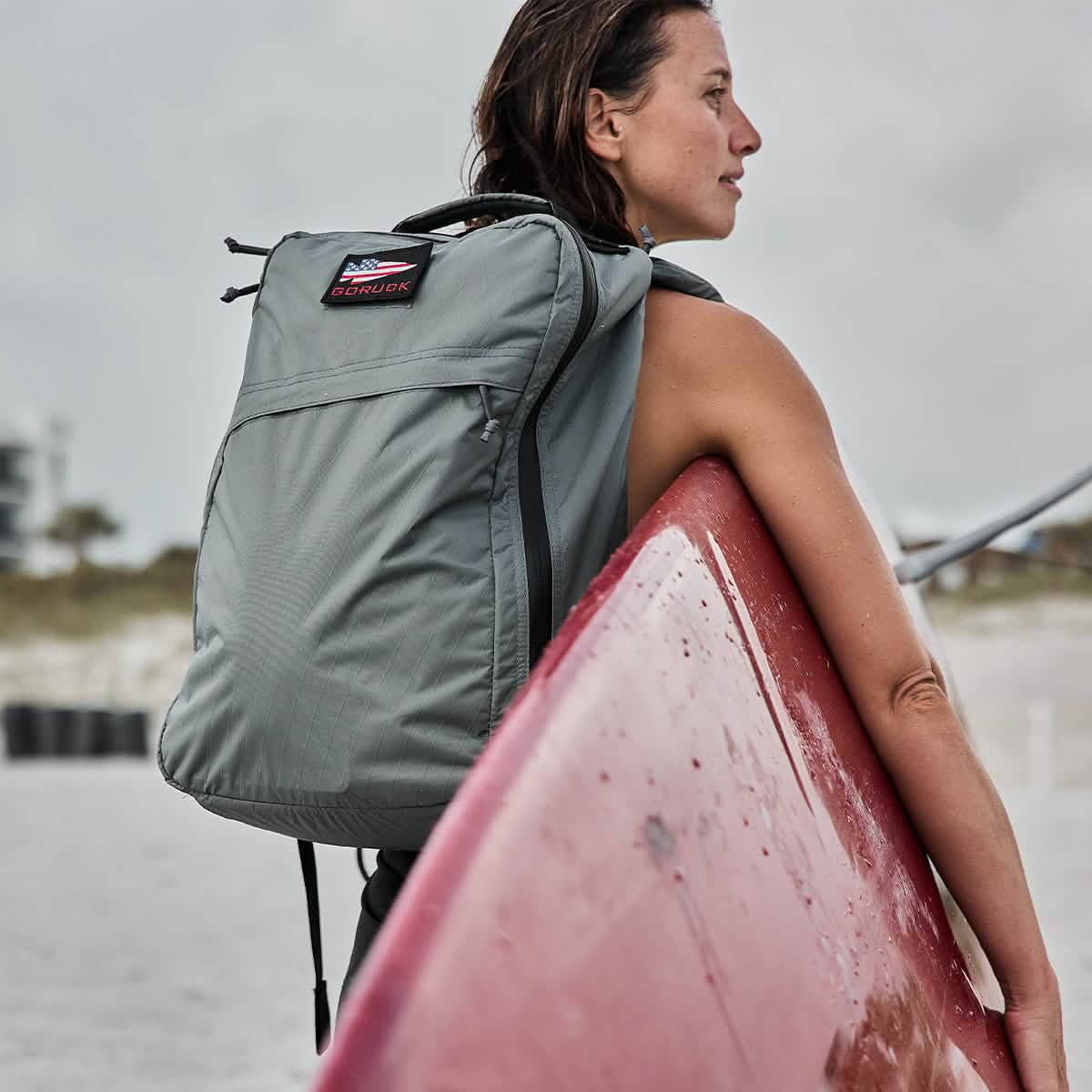 A person with short hair stands on a beach, holding a red surfboard. They carry a gray GORUCK GR1 USA - X-PAC waterproof rucksack and gaze toward the ocean under a cloudy sky, with sand dunes and sea grass in the background.