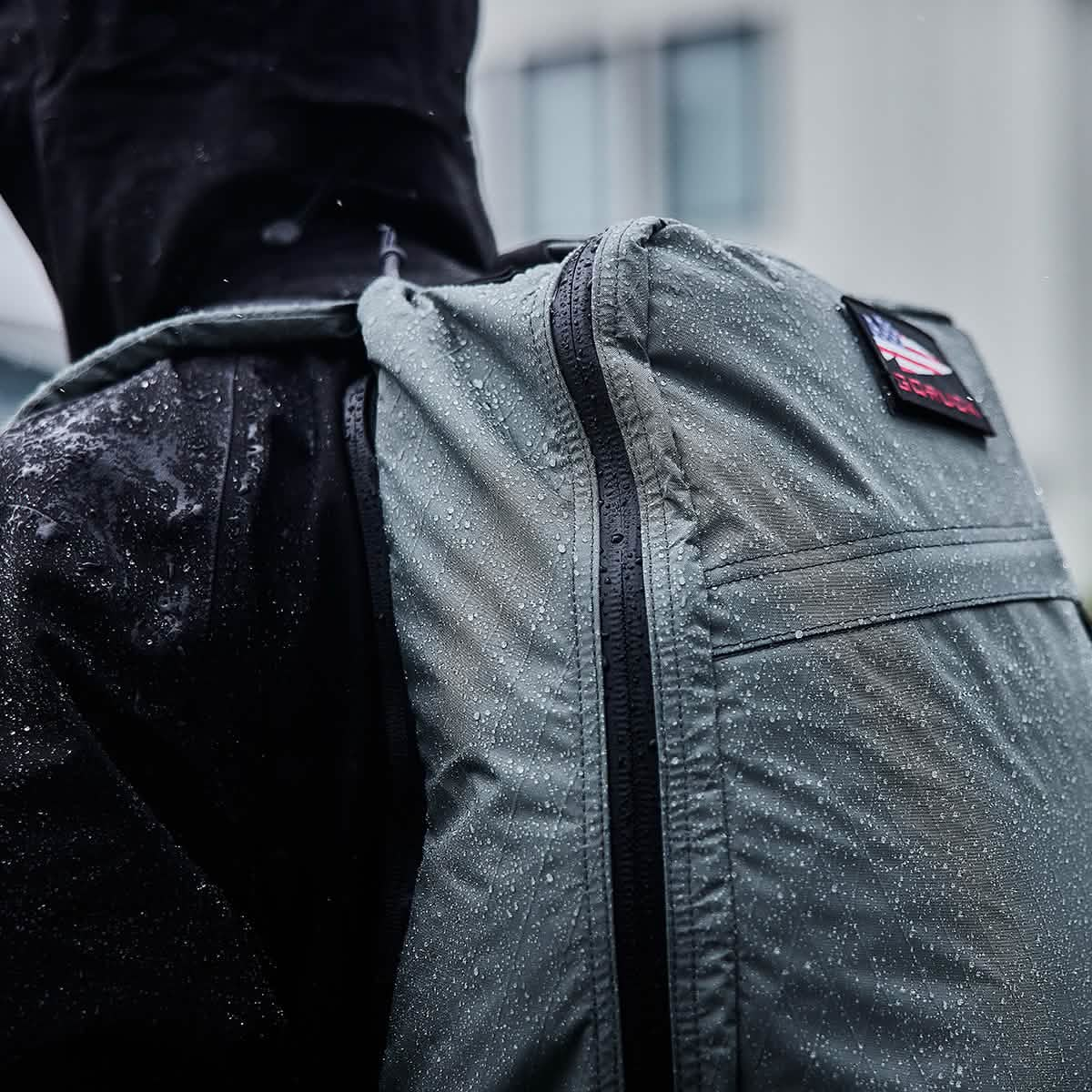 A person in a raincoat with a hood stands outdoors in the rain, their GORUCK GR1 USA - X-PAC backpack adorned with glistening water droplets.