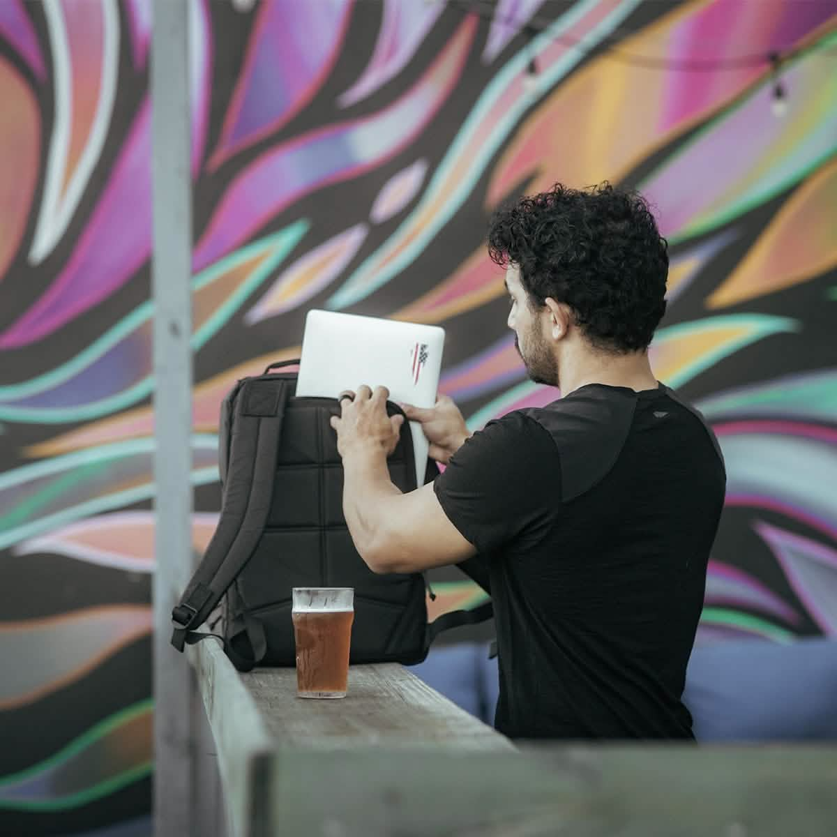 A man in a black shirt slips a laptop into a GORUCK GR1 USA - X-PAC rucksack on an outdoor wooden table. Beside the bag, there's a glass of amber-colored beer. A bright, abstract mural with swirling shapes adorns the wall behind him.