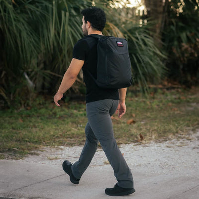 Dressed in a black shirt and gray pants, a person strolls along the sidewalk carrying a GR1 USA - X-PAC waterproof rucksack by GORUCK. They are outside, surrounded by lush greenery in the background.