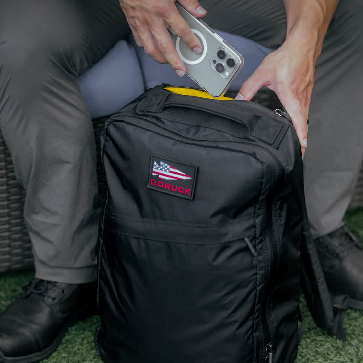 A person is sitting on a chair, holding a smartphone and a GR1 USA - X-PAC backpack by GORUCK, which features an American flag patch. They are wearing gray pants and black shoes, with grass visible beneath the chair.