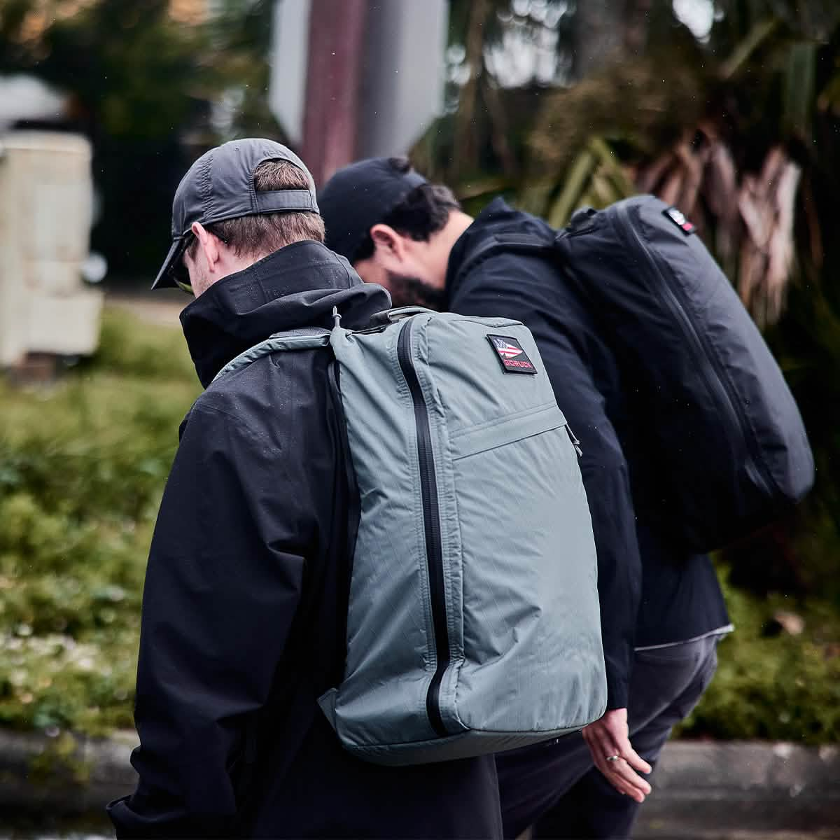 Two people with rucksacks are walking outdoors in a natural setting. One sports a cap and light-colored backpack, while the other carries a GORUCK GR1 USA - X-PAC bag in a darker shade. Casually dressed, they move along a scenic pathway, surrounded by lush greenery in the background.