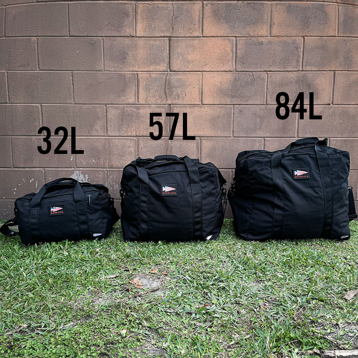 Three black deployment bags of varying sizes are arranged on grass against a brick wall backdrop. These bags echo the durability of GORUCK's Kit Bag - 84L (Includes Shoulder Strap) and are clearly marked with their capacities: 32L, 57L, and 84L, from left to right.