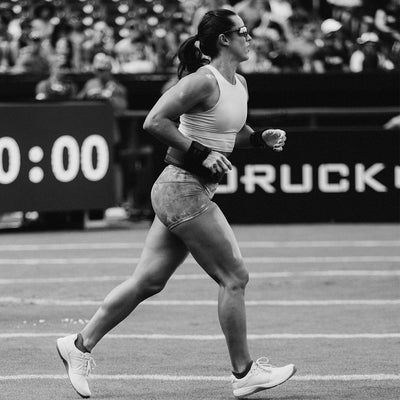 A female athlete sprints down the track, wearing a tank top, shorts, and GORUCK Women's Ballistic Trainers in Lunar Rock + Gum with a Silver Reflective Spearhead. Her long hair is tied back under her sunglasses as she maintains focus. Spectators watch in the background while a digital timer reads 0:00. The black-and-white image captures the intensity of the moment.