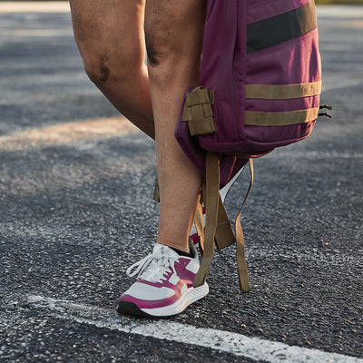 Standing outdoors on a paved surface, a person wearing the Women's Rough Runner - Baton Rouge athletic shoes by GORUCK, featuring an EVA midsole in white and pink, is holding a purple and brown backpack. Their legs are slightly bent, casting shadows on the ground.