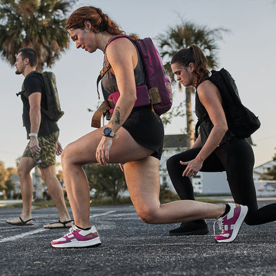 Three individuals are exercising outside. The person in the foreground lunges with a weighted vest, wearing a tank top and shorts suitable for the Women's Rough Runner - Baton Rouge by GORUCK. Nearby, another person dressed in black is kneeling on one knee, while a third individual walks in the background. The scene is beautifully set with palm trees and a clear sky.