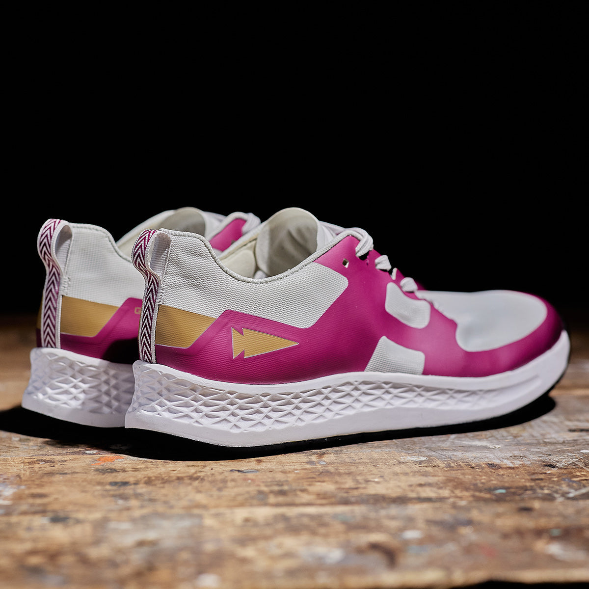 The Women's Rough Runner - Baton Rouge by GORUCK are stylish pink and white athletic shoes with gold accents, resting on a wooden surface. They showcase a geometric pattern on the EVA midsole and a zigzag design on the heel pull-tabs, making them perfect for runners who want both style and performance.