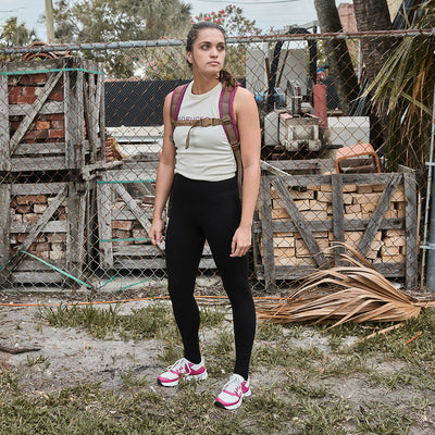 A person stands outdoors in Women's Rough Runner - Baton Rouge gear by GORUCK, wearing a light-colored sleeveless top, black leggings, and pink-trimmed sneakers featuring an EVA Midsole. They carry a backpack and are near a chain-link fence with stacked wood. Trees and a cloudy sky form the backdrop.