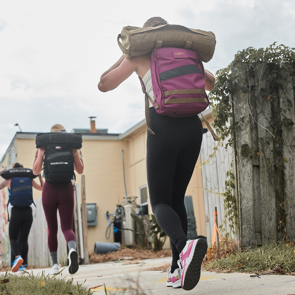 Three women wearing GORUCK Women's Rough Runner - Baton Rouge athletic clothing and backpacks jog along a path bordered by a fence and lush vegetation. One of them carries a sandbag on her shoulders, showcasing her endurance. The scene unfolds under a partly cloudy sky, enhancing the dynamic atmosphere.