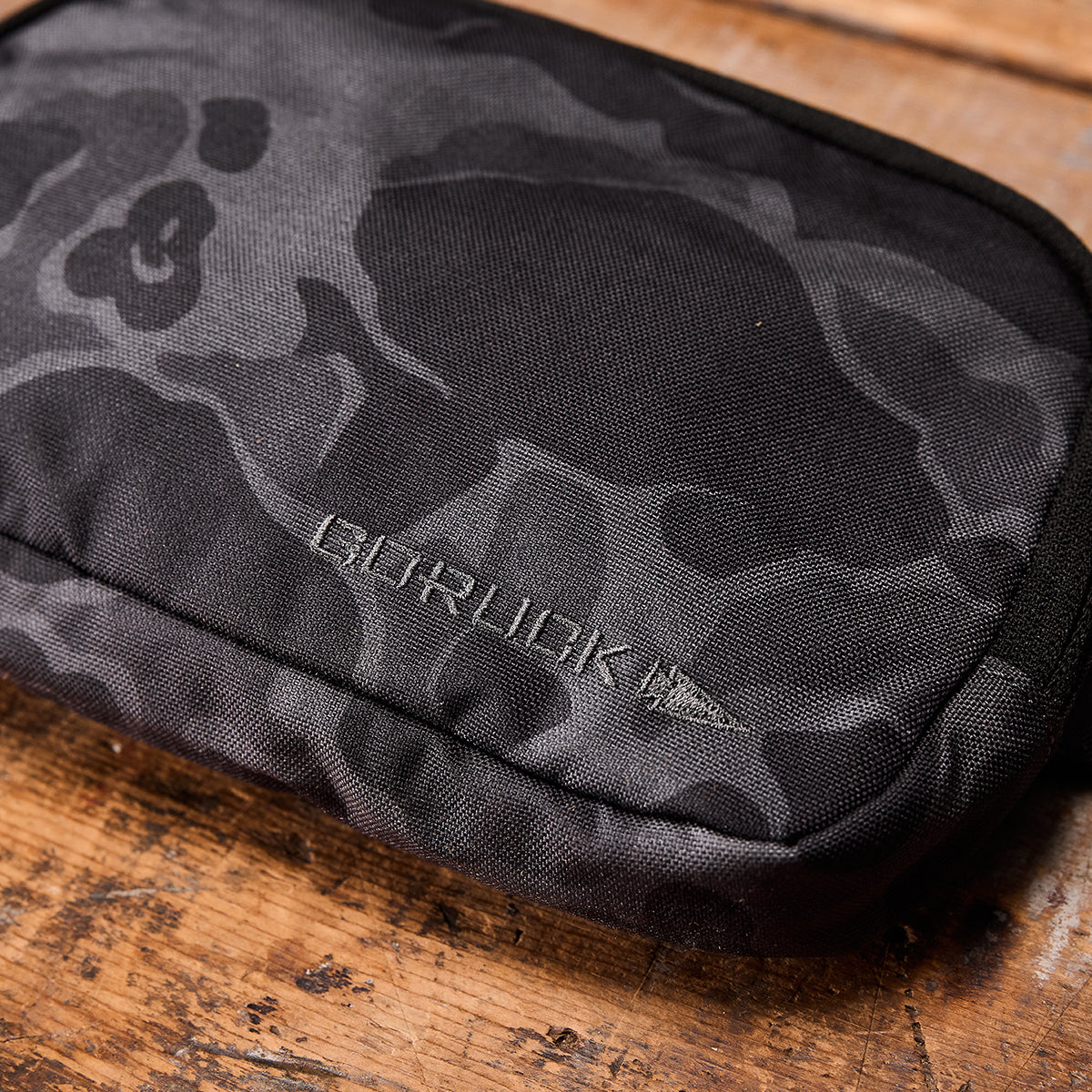 The Belt Bag by GORUCK, featuring a black and gray camo pattern and zipper, is shown resting on a wooden surface. The GORUCK name and arrowhead logo are clearly visible on the compact fabric pouch designed for belt attachment.