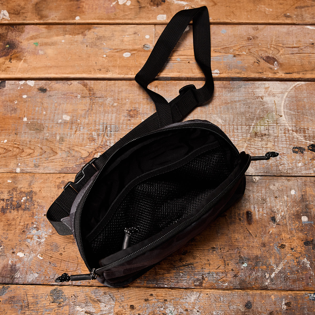 A GORUCK Belt Bag in black, featuring an adaptable strap and open zipper, rests on a rustic, paint-splattered wooden surface. The compact design includes an adjustable webbing and a mesh compartment inside.