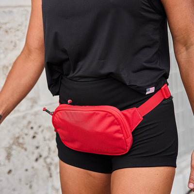 A person in a black tank top and shorts walks while highlighting a GORUCK Belt Bag with adjustable webbing. The vibrant red bag contrasts strikingly against the textured stone wall in the background.