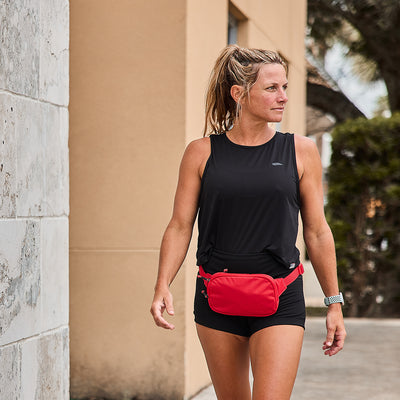 A woman in a black tank top and shorts strolls outdoors while carrying a compact Belt Bag from GORUCK in red, featuring adjustable webbing. Her hair is styled in a ponytail as she gazes to the side, with a building and trees serving as her backdrop, all while wearing her favorite watch.