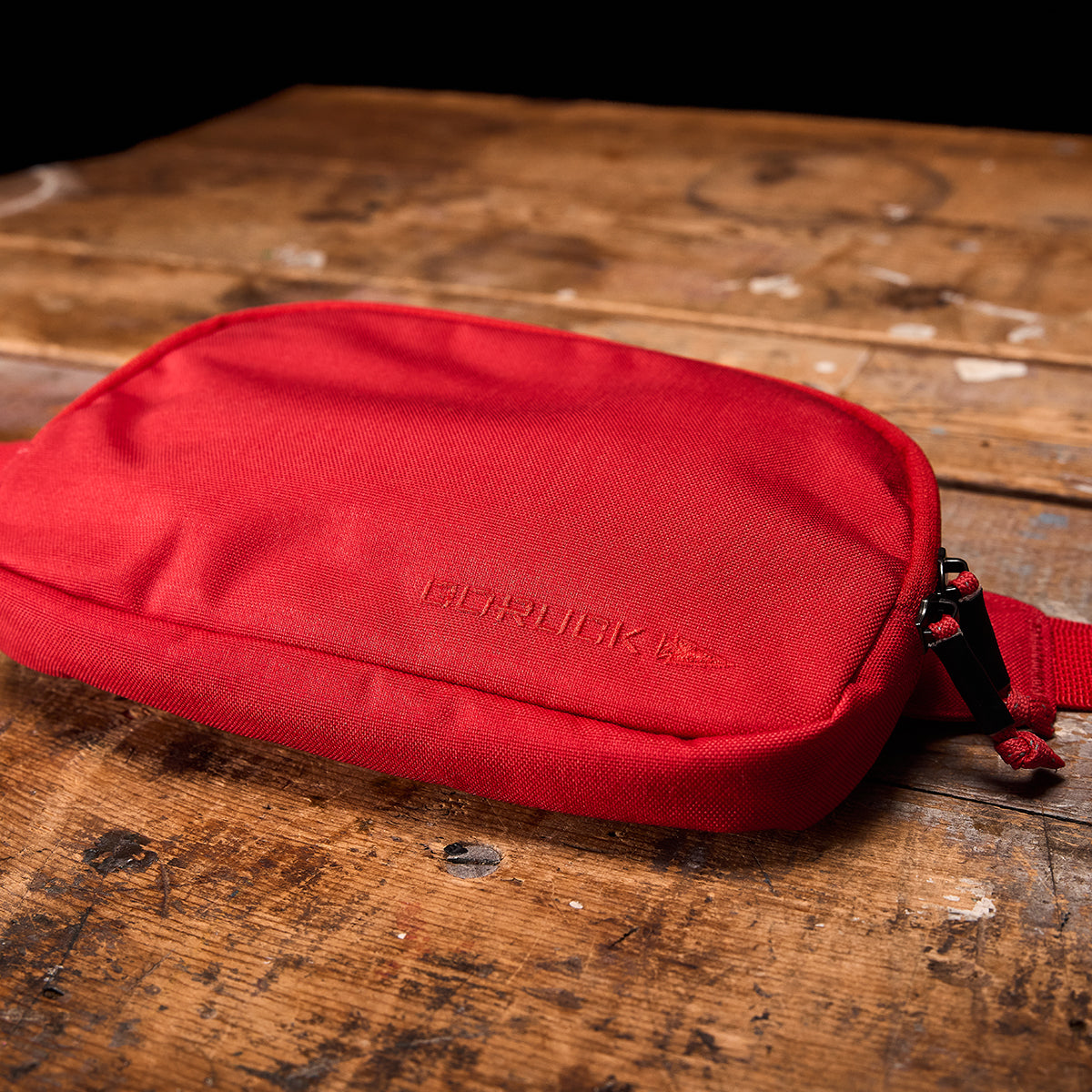 A red rectangular GORUCK Belt Bag with a zipper rests on a rustic wooden surface. The fabric boasts a textured appearance, and the brand name is subtly embossed on the front, ensuring your gear stays organized.