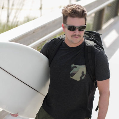 A person wearing a GORUCK x Birdwell Tee - Tri-Blend featuring a camouflage pocket, along with sunglasses and a mustache, carries a surfboard. They are strolling along the boardwalk near the beach with a GORUCK Scars backpack.