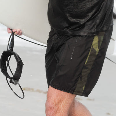 A person in 300 Boardshorts by GORUCK, designed in black and green with SurfNyl fabric and crafted in the USA, walks on the beach holding a surfboard and leash, with focus on their lower body.