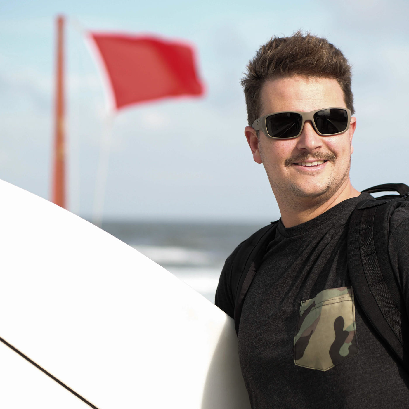 A person wearing sunglasses and a GORUCK x Birdwell Tee - Tri-Blend smiles while standing near a surfboard at the beach. A red flag flutters in the background, signaling caution. The sky is partly cloudy, highlighting the charm of American craftsmanship evident all around.