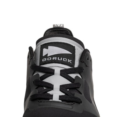 Close-up of the Women's Rough Runner - Black + White sneaker featuring a black and gray design with black laces. The label on the tongue reads GORUCK. This shoe, inspired by Special Forces, showcases a blend of matte and glossy textures with geometric shapes, making it perfect for the Rough Runner™ in you.