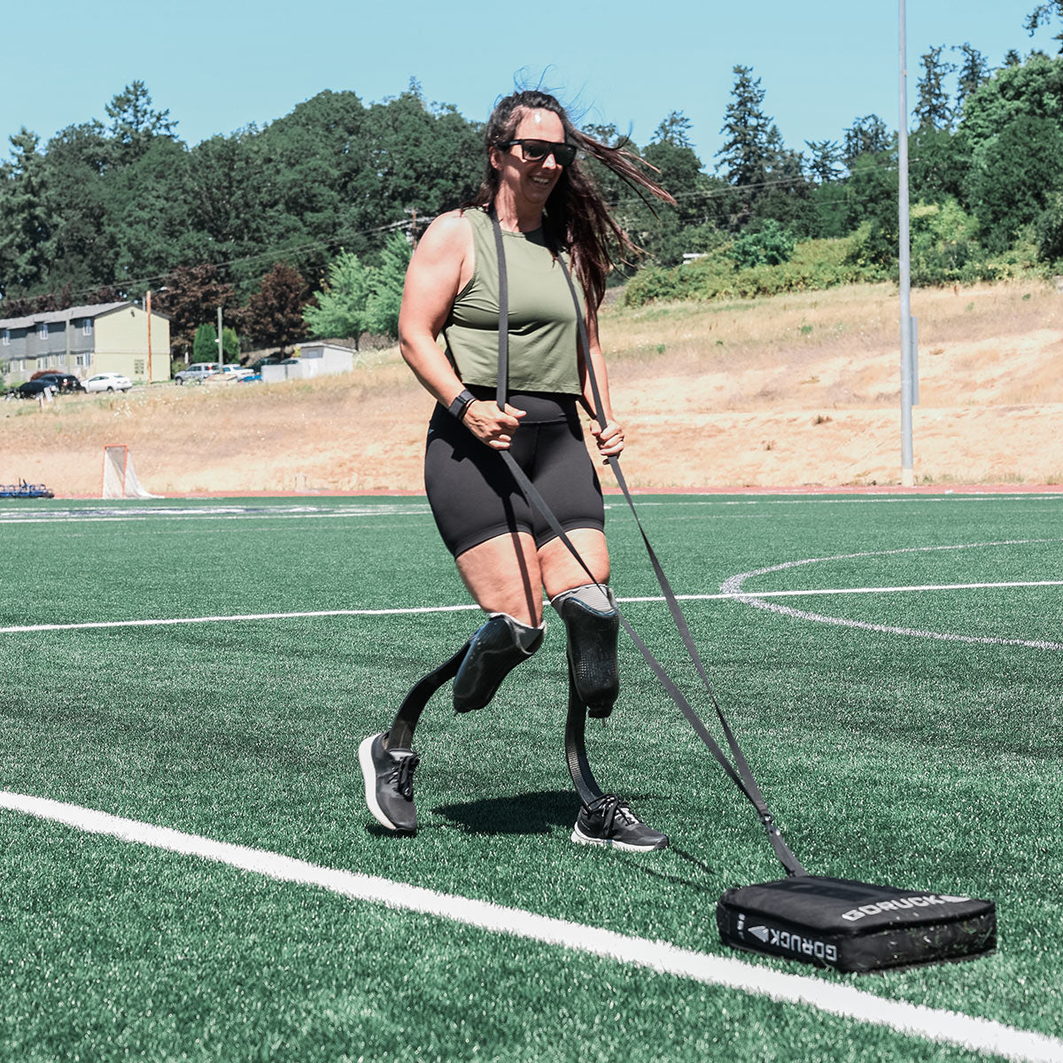 A woman with prosthetic legs, capturing the essence of GORUCK's Women's Rough Runner - Black + White, exercises on a track field while pulling a weighted sled. She sports a green tank top, black shorts, and sunglasses against the backdrop of trees and a building.