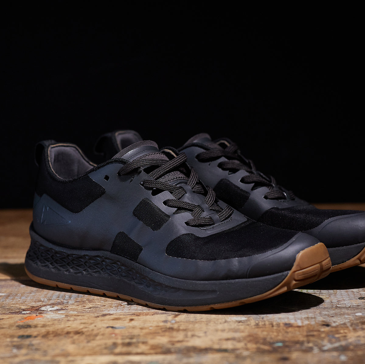 The Women's Rough Runner - Black + Gum by GORUCK, featuring black laces and various fabric textures, are displayed with textured soles and brown rubber accents on the toes. The shoes boast an EVA midsole for added comfort, positioned on a wooden surface against a dark background.