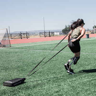 A woman with a prosthetic leg is charging through a grassy field with the determination of Special Forces, pulling a sled behind her. Wearing the Women's Rough Runner - Black + White from GORUCK, and sunglasses, her hair tied back, she stands out against the track and open sky backdrop.