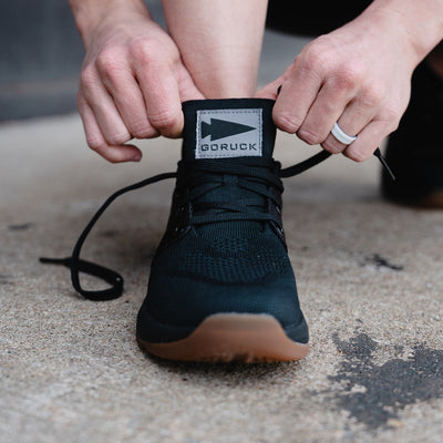 An individual ties the laces of a Women's Ballistic Trainer from GORUCK, designed for functional fitness, while on a concrete surface. The shoe showcases a gum sole and is crafted from CORDURA® Ballistic Nylon with a visible fabric label on the tongue. The person wears a light-colored ring and concentrates on their workout footwear.