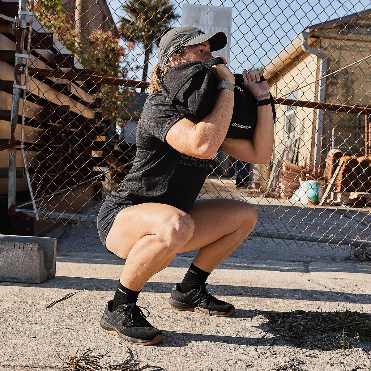 Wearing GORUCK Women's Ballistic Trainers in Black + Gum with a Black Reflective Spearhead, a person performs a squat exercise outdoors, holding a sandbag close to their chest. They are positioned in front of a chain-link fence, with buildings and plants visible in the background under a clear blue sky.