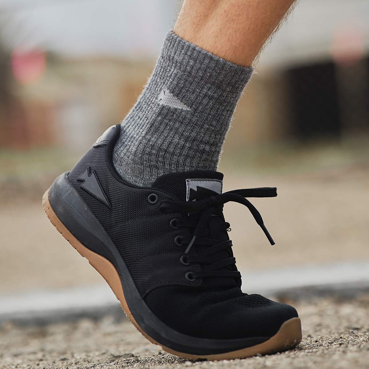 A person in gray socks is stepping on a gravel path, wearing GORUCK Women's Ballistic Trainers in Black + Gum with a Black Reflective Spearhead. These functional fitness shoes are crafted from durable CORDURA® Ballistic Nylon and feature a subtle arrow logo on the side. The background is blurred, emphasizing the footwear.