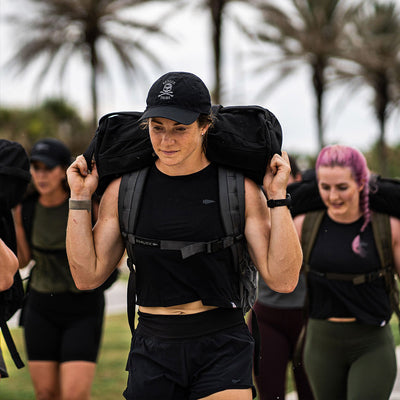 A group of people are exercising outdoors, each carrying a large backpack. They walk in a line under palm trees, with the focus on a person wearing the Women’s USA Performance Tank - ToughMesh and a black cap. This tank, by GORUCK, is crafted from ToughMesh™ fabric that dries lightning fast, enhancing their determined and energetic atmosphere.