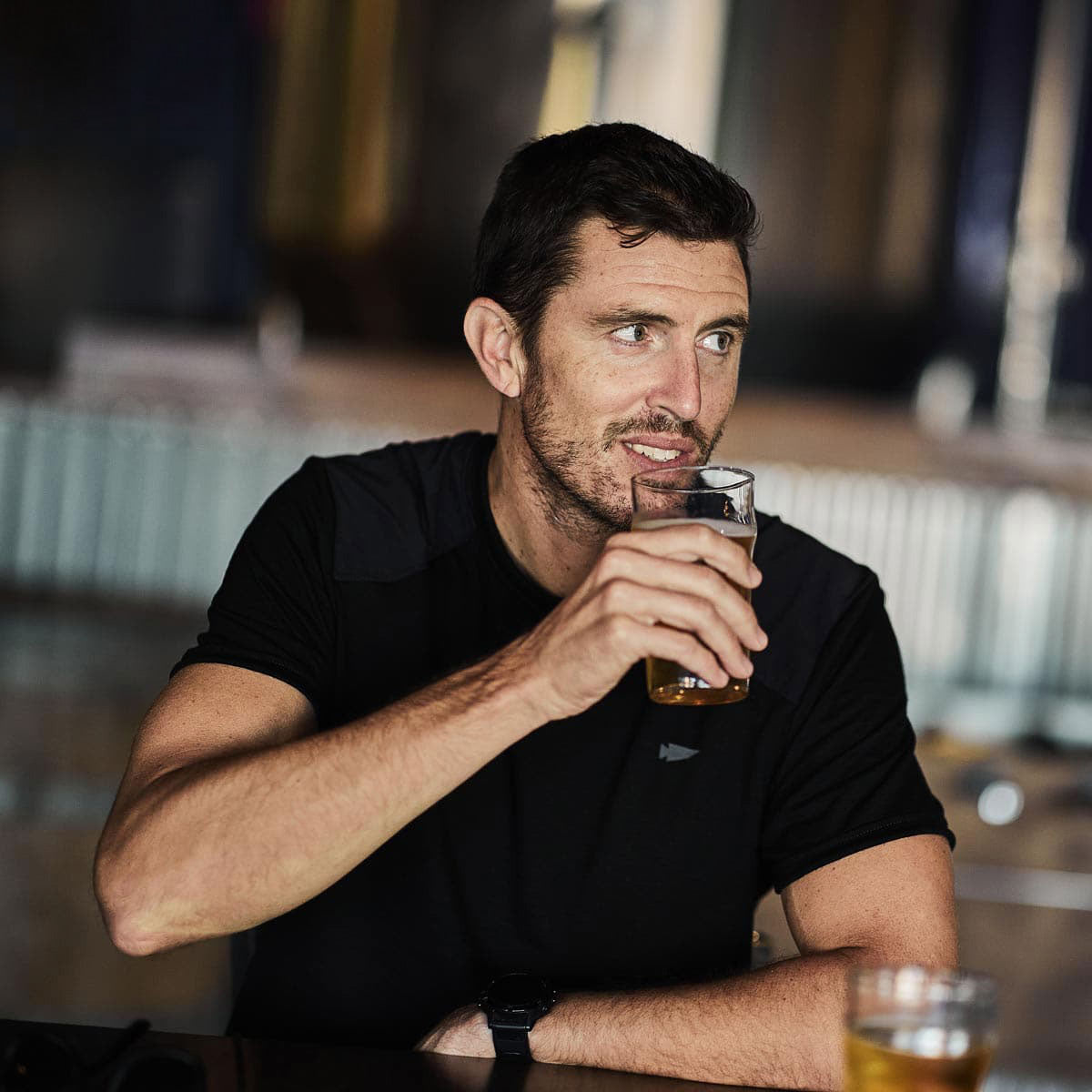 A man wearing the GORUCK Men's Commando Tee in merino wool sits at a table holding a glass. He gazes to the side, seemingly enveloped in the cozy ambiance of a casual setting, with warm lighting softening the blurry background.