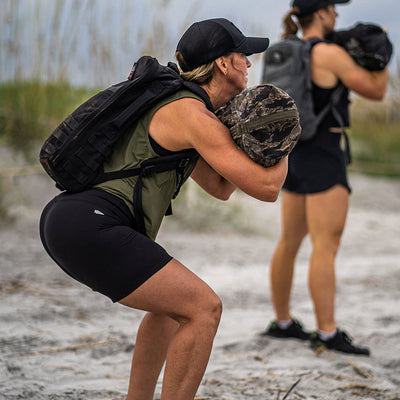 Two individuals wearing backpacks and caps perform squats on a sandy beach, holding weighted sacks. They are dressed in athletic gear featuring GORUCK's Women’s Biker Shorts - ToughFlex, with grasses visible in the background.