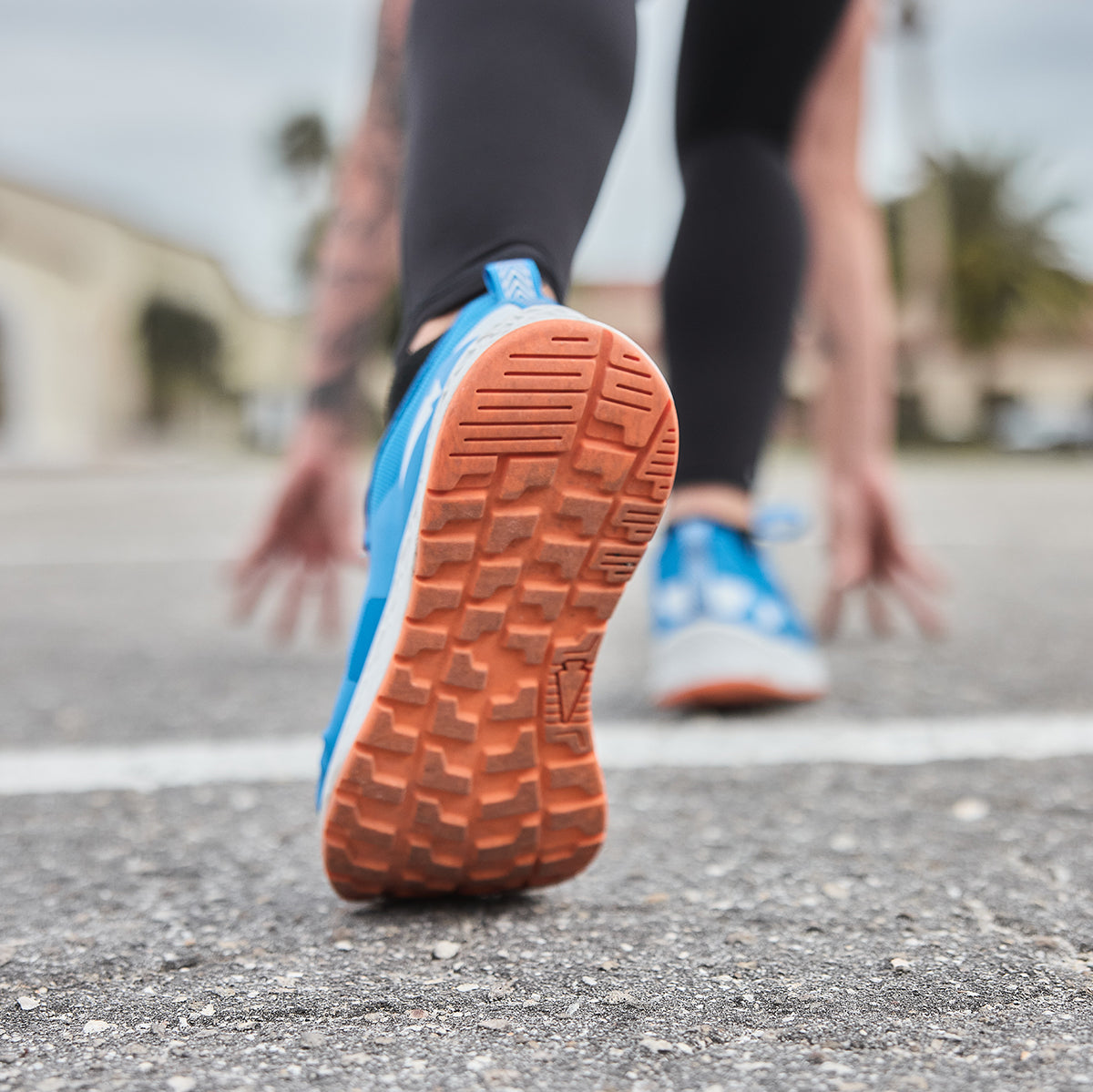 A close-up captures a runner in the Women's Rough Runner - Electric Blue by GORUCK, poised to start with the vibrant blue and orange sole of her shoe firmly on the pavement. Her fingertips graze the road as buildings and trees blur in the backdrop, embodying a spirit built for high mileage.