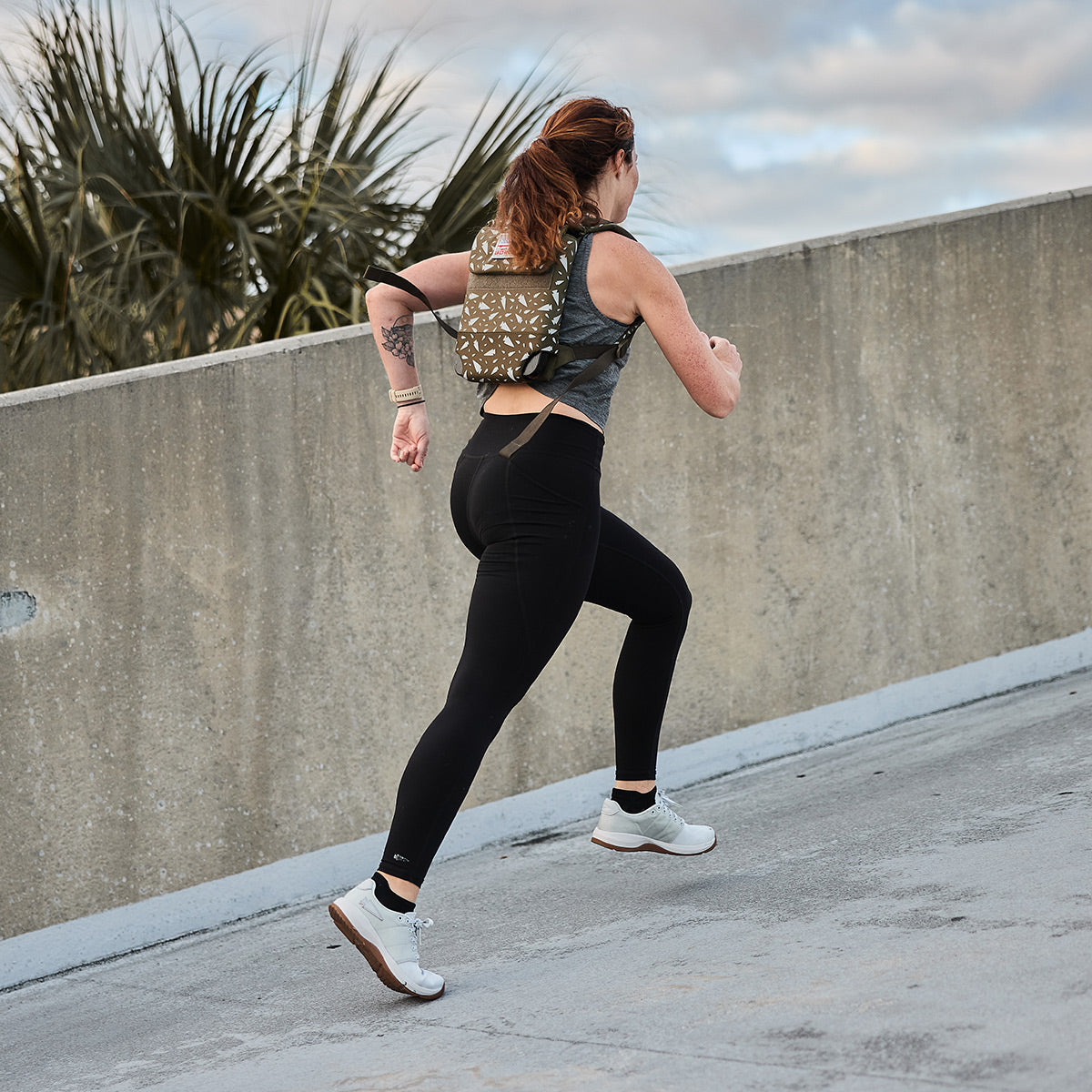 A person with long hair runs uphill on a concrete path, wearing the Women's Ballistic Trainers - Lunar Rock + Gum with a Silver Reflective Spearhead from GORUCK. They have on a backpack, a dark tank top, and black leggings. Palm trees and a cloudy sky create the perfect backdrop for their invigorating workout in those functional fitness shoes.