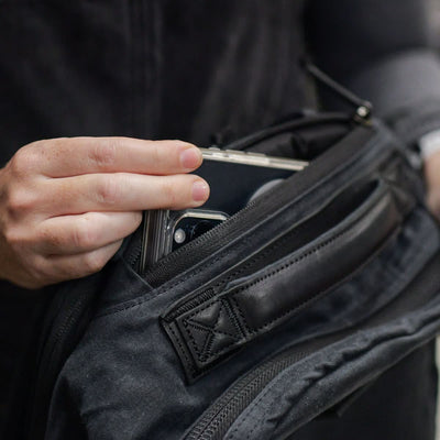 A person is placing a smartphone into a small, black Bullet Ruck made from waxed canvas by GORUCK. The image highlights the hands and the bag, drawing attention to the phone's camera lenses. The person's dark clothing complements the sleek, retro look of the GORUCK canvas rucksack.