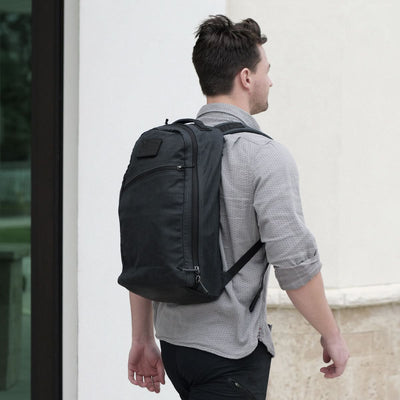 A person with short dark hair is strolling outdoors, wearing a grey button-up shirt and carrying the Bullet Ruck - Waxed Canvas by GORUCK. As they pass by a window and a light-colored wall, their retro GORUCK canvas rucksack adds a touch of timeless style to their look.