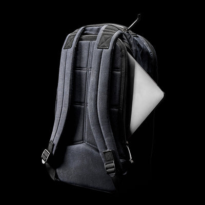 Against a dark backdrop, the GORUCK Bullet Ruck - Waxed Canvas in sleek black showcases its padded shoulder straps and ergonomic back support, while a silver laptop partially visible from the top pocket exudes modern utility combined with timeless style.