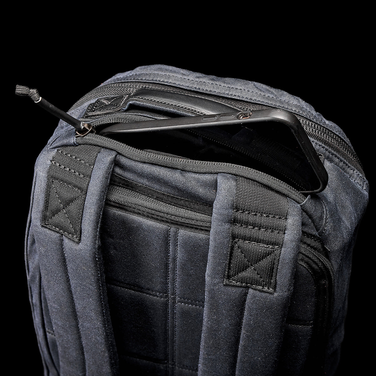 A close-up of the GORUCK Bullet Ruck - Waxed Canvas against a dark background. The top compartment is slightly open, revealing a smartphone inside with a small zipper partially closed. The backpack features quilted stitching and sturdy straps, showcasing its rugged charm.