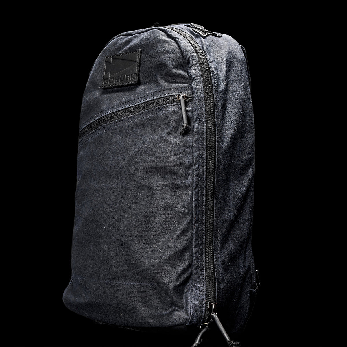 A GORUCK Bullet Ruck - Waxed Canvas backpack in black, showcasing a sleek and minimalist design, is presented against a dark backdrop. It includes a zippered front pocket and features a subtle logo near the top.