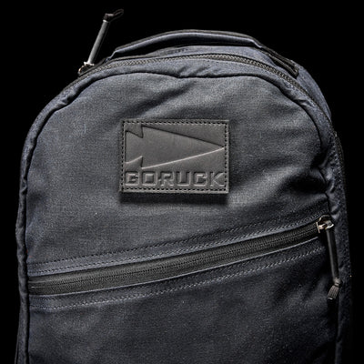 The Bullet Ruck - Waxed Canvas backpack by GORUCK is displayed. Its dark grey waxed canvas construction includes a zippered front pocket, and it features the GORUCK logo on a fabric patch, set against a black background.