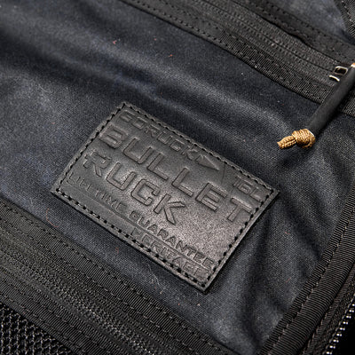 Close-up of a black fabric bag featuring a stitched leather patch with embossed text: Bullet Ruck and Lifetime Guarantee by GORUCK. The background shows part of a zipper with a corded pull tab, enhancing the rugged appeal reminiscent of the Bullet Ruck - Waxed Canvas.