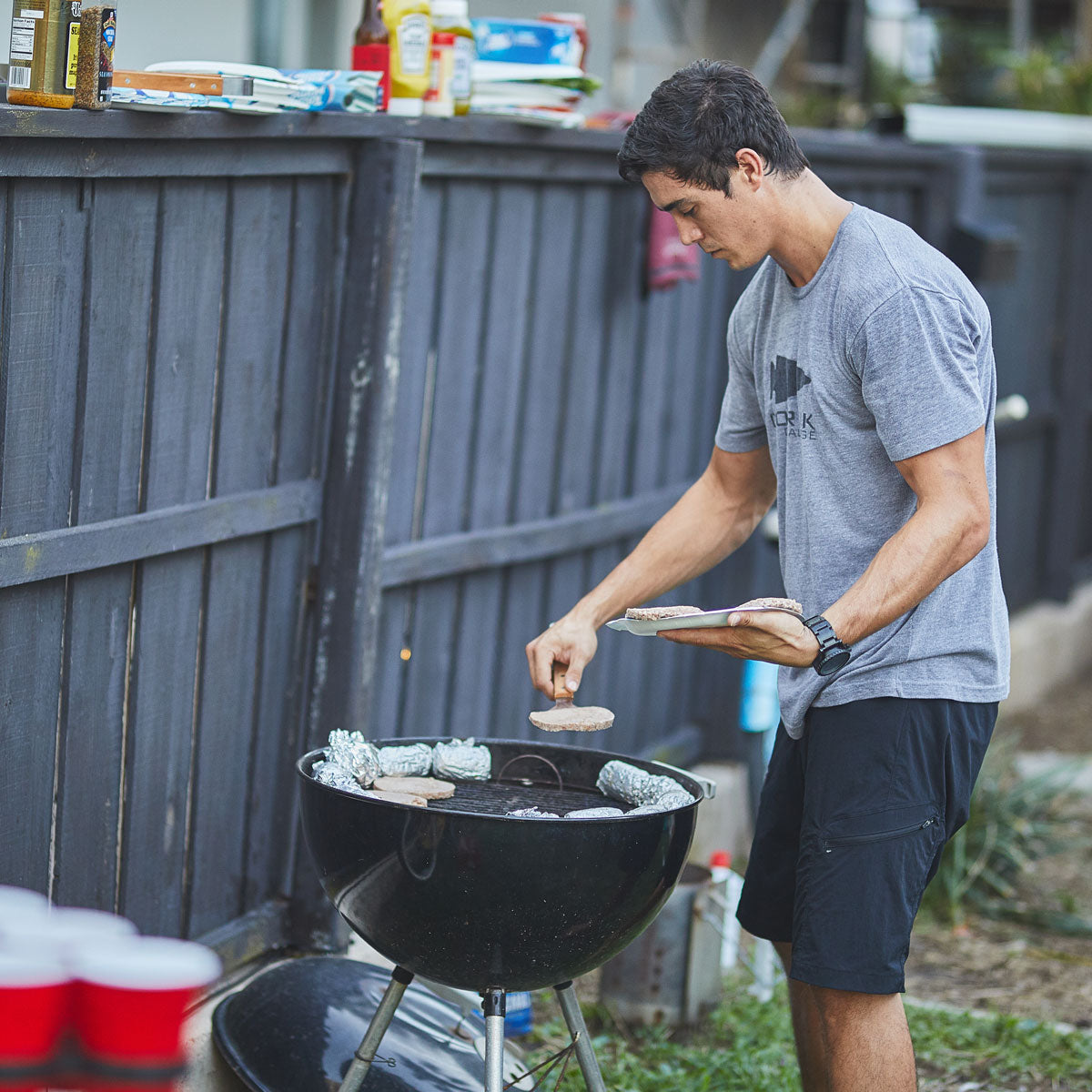 A man wearing GORUCK's Men’s Challenge Shorts in black, made with lightweight ToughDry® fabric, is grilling burgers on a charcoal barbecue grill outdoors. He is holding a spatula and a wooden board with patties, while red cups and condiments are set on a nearby table by a wooden fence.