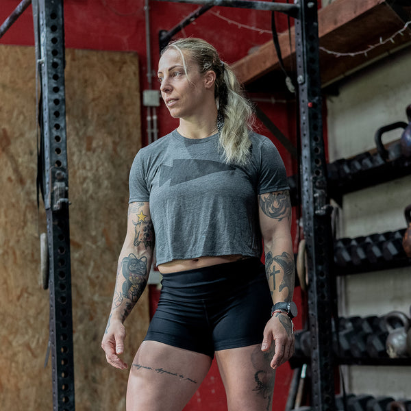 A tattooed individual stands by gym equipment, wearing a Women's Cropped Tee - Poly-Blend in gray and black shorts, glancing sideways.