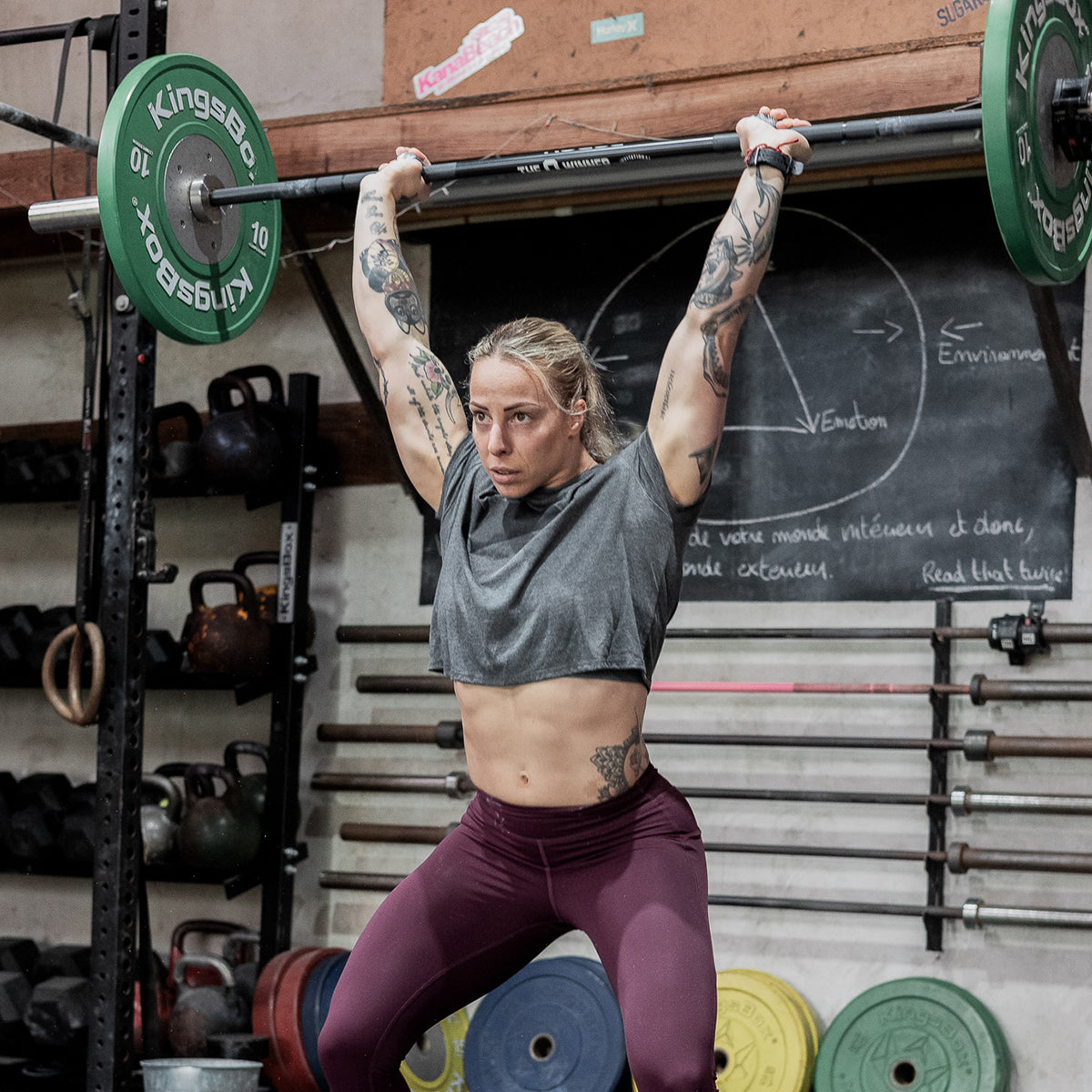 A person lifting a barbell overhead in a gym, wearing a Women's Cropped Tee - Poly-Blend in gray and maroon leggings, surrounded by workout equipment.